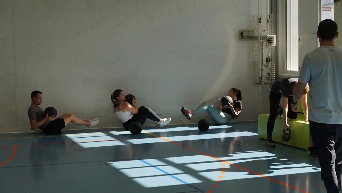 Man sieht Teilnemer am Boden sitzen mit Gewichten,  und führen mit diesen eine Übung aus. 